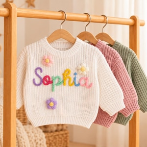 May include: Three long-sleeve knit sweaters hanging on wooden hangers. The front white sweater features the name Sophia in colorful raised yarn lettering, accented with three small, dimensional yarn flowers in pink, white, and purple.