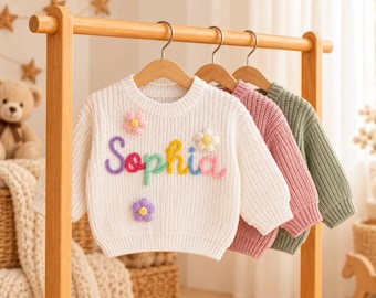 May include: Three long-sleeve knit sweaters hanging on wooden hangers. The front white sweater features the name Sophia in colorful raised yarn lettering, accented with three small, dimensional yarn flowers in pink, white, and purple.