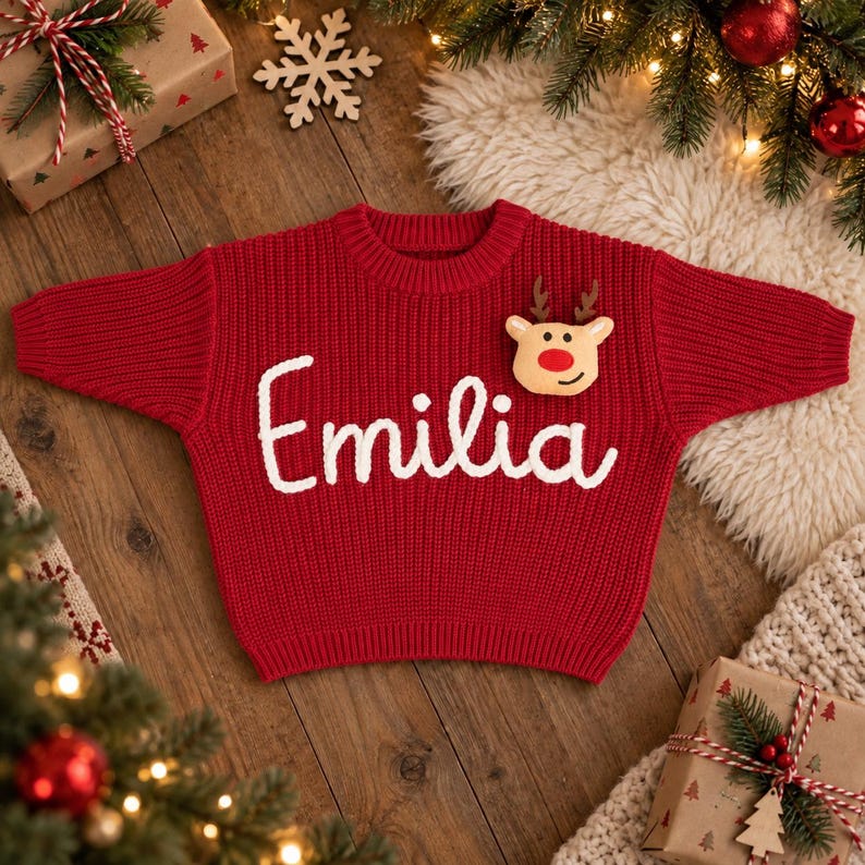 May include: A red long-sleeve knit sweater featuring the name Emilia embroidered in white script across the chest. A plush reindeer head with brown antlers and a bright red nose is attached to the upper right side of the sweater front.