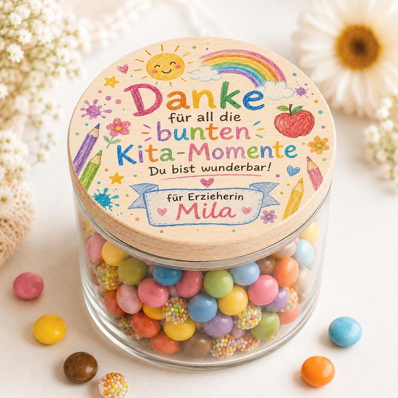 May include: A clear glass jar filled with colorful round candies, topped with a wooden lid featuring a printed design of a sun, rainbow, apple, and pencils. The text reads: Danke für all die bunten Kita-Momente Du bist wunderbar! für Erzieherin Mila.