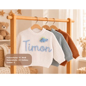 May include: A white long-sleeve knit sweater featuring the name Timon embroidered in light blue script and a small light blue dinosaur appliqu&eacute;. The sweater hangs on a wooden hanger alongside two other knit sweaters in blue and rust colors.