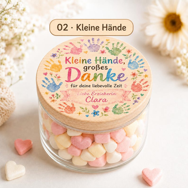 Puede incluir: Un frasco de vidrio transparente lleno de dulces en forma de coraz&oacute;n, con una tapa de madera que presenta ilustraciones de huellas de manos coloridas, motivos florales y el texto en alem&aacute;n "Kleine H&auml;nde, gro&szlig;es Danke f&uuml;r deine liebevolle Zeit" y "Clara".
