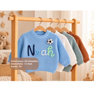 May include: A light blue knit sweater featuring the name Noah in dark blue, white, and green yarn lettering, with a small soccer ball patch. The text on the image reads: Pulloverfarbe: 08 Himmelblau, Namensfarbe: 16 Noah, Symbol: ZA.