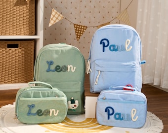 May include: Two corduroy backpacks and two matching lunch bags in light blue and sage green. Each item is personalized with embroidered names Leon or Paul in a gradient blue font. The bags feature front zippered pockets and white zipper pulls.