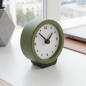 Puede incluir: Un reloj analógico verde oliva con esfera blanca y manecillas negras. El reloj está sobre una base negra y muestra la hora. El reloj está sobre un escritorio blanco con una ventana al fondo.
