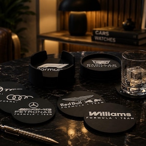 Könnte beinhalten: Ein Set aus schwarzen, runden Untersetzern mit weißen aufgedruckten Logos verschiedener Formel-1-Teams, darunter Mercedes-AMG Petronas, Red Bull, Williams, Audi und Cadillac. Die Untersetzer sind auf einer dunklen Marmoroberfläche angeordnet.
