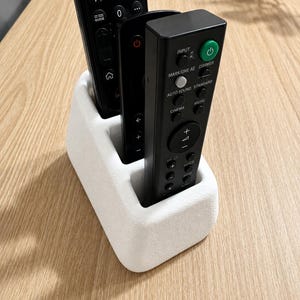 May include: A white, rectangular remote control holder with three vertical slots, currently containing three black remote controls. The front-most remote features a green power button and labels including Input, Mark/One AE, Auto Sound, and Cinema.