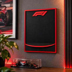 May include: A rectangular wall-mounted display case featuring a black back panel, a red frame, and a red F1 logo at the top. The unit includes a curved red shelf at the bottom, designed for showcasing collectibles in a sleek, modern presentation.