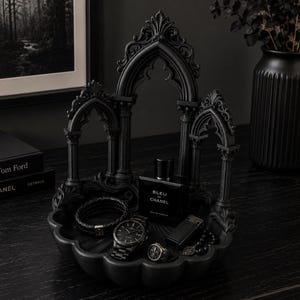 Puede incluir: Una bandeja decorativa de color negro mate con una intrincada estructura de arco gótico con columnas y detalles ornamentados. La bandeja contiene un frasco de perfume cuadrado Bleu de Chanel, un reloj de pulsera negro, una pulsera trenzada, un anillo y un pequeño objeto rectangular.