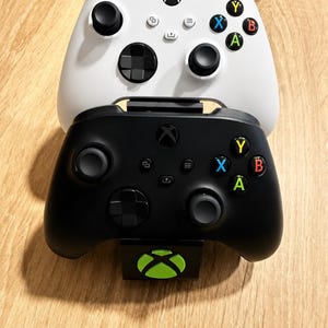 Op de afbeelding: Een witte en een zwarte draadloze gamecontroller verticaal gestapeld op een zwarte standaard. Elke controller is voorzien van twee analoge sticks, een D-pad en vier gekleurde actieknoppen met de letters Y, B, A en X. De standaard toont een groen Xbox-logo.