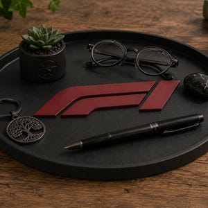 Op de afbeelding: Een rond zwart dienblad met een donkerrood Formule 1-logo, een zwarte balpen, een bril met zwart montuur, een kleine vetplant in een donkere pot, een ronde sleutelhanger met een boommotief en een kleine zwarte steen met marmerpatroon.