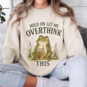 May include: A cream-colored crewneck sweatshirt featuring a graphic of a green frog holding a glass of iced coffee. The text above and below the frog reads, "HOLD ON LET ME OVERTHINK THIS" in dark green, distressed lettering, surrounded by cattails.