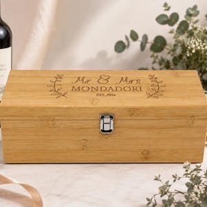May include: A rectangular bamboo wine box with a silver metal latch. The lid is engraved with the text Mr & Mrs MONDADORI EST. 2026, flanked by two decorative leaf branch designs. The box features a natural light brown wood grain finish.