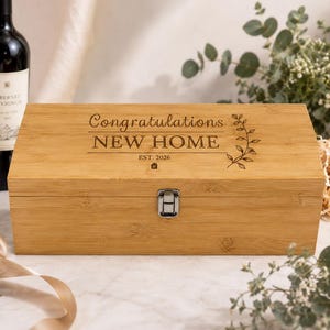 May include: A rectangular light brown bamboo wine box with a silver metal latch. The lid is engraved with the text Congratulations NEW HOME EST. 2026 and a small vine illustration. The box features a smooth wood grain finish and a hinged lid.