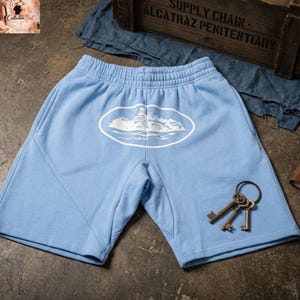 Op de afbeelding: Lichtblauwe shorts met een witte ovale afbeelding van een gebouw en water. De shorts hebben zakken en een elastische tailleband. Een set sleutels ligt in de buurt van de shorts. De shorts zijn gemaakt van een zachte stof.