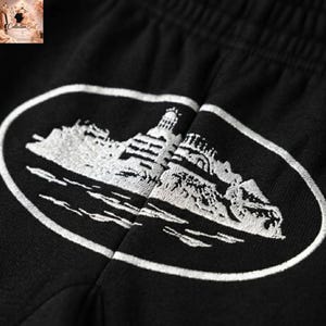 May include: Black shorts with a white embroidered design of a lighthouse on a rocky island, framed by a white oval. The shorts are made of a soft, dark fabric.