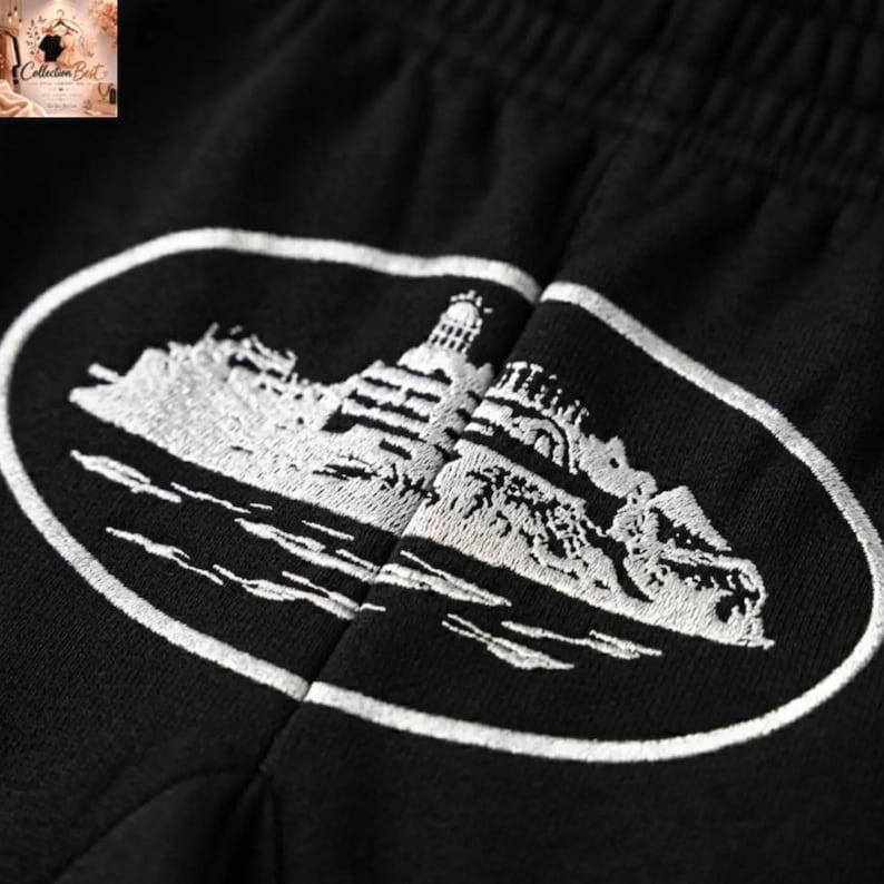 May include: Black shorts with a white embroidered design of a lighthouse on a rocky island, framed by a white oval. The shorts are made of a soft, dark fabric.