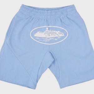 Op de afbeelding: Lichtblauwe shorts met een elastische tailleband en zijzakken. Een witte ovale afbeelding van een gebouw op water is gecentreerd op de achterkant. De shorts zijn gemaakt van een zacht materiaal.