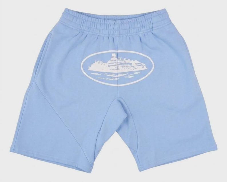Op de afbeelding: Lichtblauwe shorts met een elastische tailleband en zijzakken. Een witte ovale afbeelding van een gebouw op water is gecentreerd op de achterkant. De shorts zijn gemaakt van een zacht materiaal.