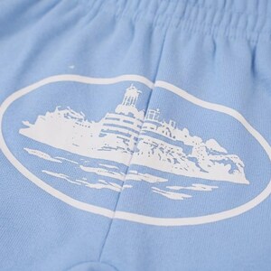 Op de afbeelding: Lichtblauwe shorts met een witte ovale afbeelding van een vuurtoren en een kustlandschap. De shorts hebben een elastische tailleband en zijn gemaakt van een zachte stof. De afbeelding staat in het midden.