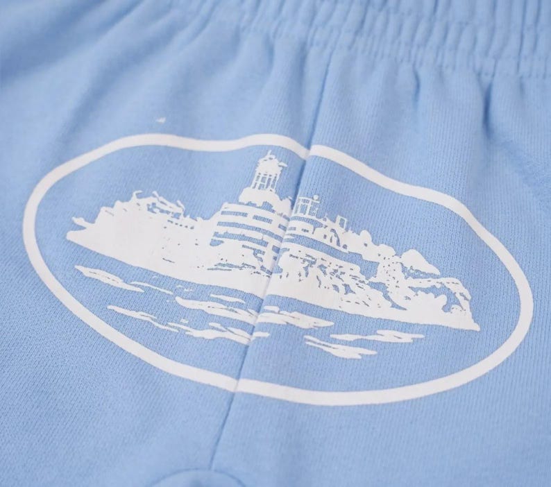 Op de afbeelding: Lichtblauwe shorts met een witte ovale afbeelding van een vuurtoren en een kustlandschap. De shorts hebben een elastische tailleband en zijn gemaakt van een zachte stof. De afbeelding staat in het midden.