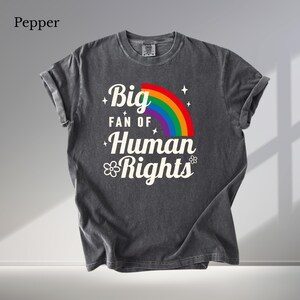 Könnte beinhalten: Ein anthrazitfarbenes Kurzarm-T-Shirt mit dem weißen Schriftzug Big FAN OF Human Rights, einem bunten Regenbogenbogen sowie kleinen Stern- und Blumenakzenten. Das Shirt hat einen Rundhalsausschnitt und eine verwaschene Vintage-Stoffoptik.