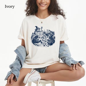May include: An ivory short-sleeved t-shirt featuring a dark blue graphic print of four cats wearing wizard hats and robes while playing a tabletop role-playing game with dice and a game board. The text Ivory is printed in the top left corner.