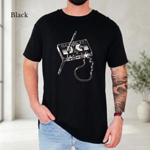 Può includere: Una t-shirt nera a maniche corte con una stampa grafica bianca di una cassetta audio con una matita inserita in una delle bobine. La cassetta riporta la scritta "OLD SCHOOL I.T." con un nastro magnetico che pende verso il basso.