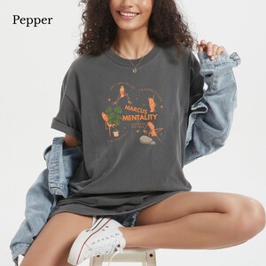May include: A charcoal gray t-shirt featuring a graphic print with orange illustrations of insects and the text Marcus Mentality, don't piss me off, I'm mildly irritated, I'm sensitive, and I don't like this rock, paired with a light blue denim jacket.