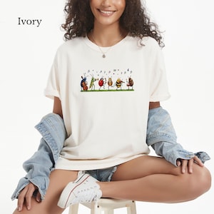 May include: An ivory t-shirt featuring a graphic print of six colorful insects playing musical instruments, including a beetle with a cello, a grasshopper with a trumpet, a ladybug with maracas, a bee with a violin, and a bug playing drums.