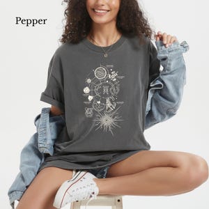 May include: A charcoal gray short-sleeve t-shirt featuring a white graphic print of various celestial bodies, geometric lines, and small text labels arranged in a circular pattern, with a large starburst design at the bottom of the graphic.