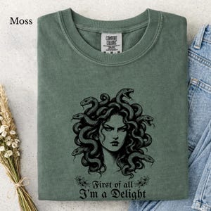 Könnte beinhalten: Ein moosgrünes T-Shirt mit einem schwarzen Grafikdruck von Medusa mit Schlangen als Haaren. Unter der Abbildung steht der Text, First of all I'm a Delight. Das T-Shirt hat einen Rundhalsausschnitt und ein Comfort Colors Etikett am Innenkragen.