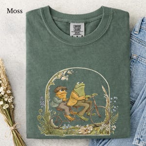 Könnte beinhalten: Ein moosgrünes T-Shirt mit Rundhalsausschnitt und einer grafischen Illustration von zwei Fröschen auf einem Tandemfahrrad. Die Frösche tragen Jacken und Hüte im Vintage-Stil und sind von einem Blumenbogen umgeben. Comfort Colors Etikett.