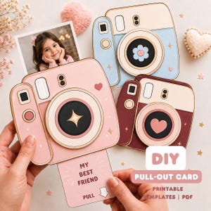 May include: Three camera-shaped paper cards in light blue, pink, and dark red. The pink card has a pull-out section with the text 'MY BEST FRIEND' and 'PULL' with an arrow. Each card features a lens with a central icon, including a flower, star, and heart.