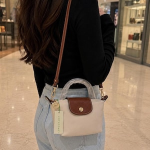 May include: A cream-colored handbag with a brown flap and handle is shown. The bag has a long, brown strap and gold-colored hardware. A tag with the word "LONGCHAMP" is attached to the bag.