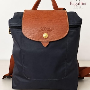 Puede incluir: Mochila azul marino con solapa y asa de cuero marrón. La solapa se asegura con un botón dorado. La bolsa tiene correas marrones y cierre de cremallera. La marca "Bagallini" es visible.