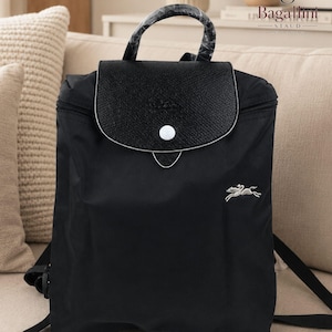 Puede incluir: Mochila negra con cierre de solapa y asa superior. La solapa es de material negro texturizado con cierre de botón blanco. Un emblema plateado de un caballo corriendo está en la parte delantera.