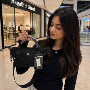 May include: A black handbag with a top handle and a detachable shoulder strap. The bag has a flap closure with a silver-tone button. A tag is attached to the handle. The store name "Bagallini Staud" is visible in the background.