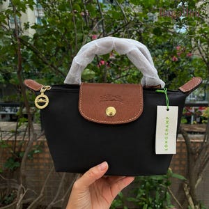 May include: Black handbag with a brown flap and handle. The bag has a gold zipper and a gold button on the flap. A white tag with the word "LONGCHAMP" is attached to the bag.