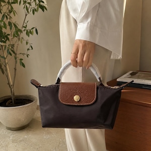 May include: A dark brown handbag with a brown leather flap and a gold-colored button closure. The bag has a white handle and a zipper closure. The bag is held by a person wearing a white shirt.