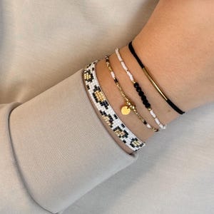 Pode incluir: Um conjunto de quatro pulseiras com uma banda larga de missangas com padrão de leopardo preto, branco e dourado, uma corrente fina dourada com um pequeno pendente redondo, um fio de missangas brancas com detalhes pretos e um cordão preto com tubo dourado.