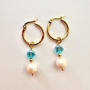Pode incluir: Um par de brincos de argola dourados com pingentes pendentes. Cada brinco consiste numa pequena argola dourada, uma conta facetada azul-clara translúcida, uma pequena conta espaçadora dourada e uma conta branca perolada na ponta.