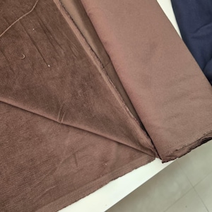 Pu&ograve; includere: Un rotolo di tessuto in velluto a coste marrone caratterizzato da una distinta trama a coste verticali. Il materiale &egrave; ripiegato per rivelare la superficie morbida e opaca del tessuto, mostrando il colore marrone profondo e la trama tessile.