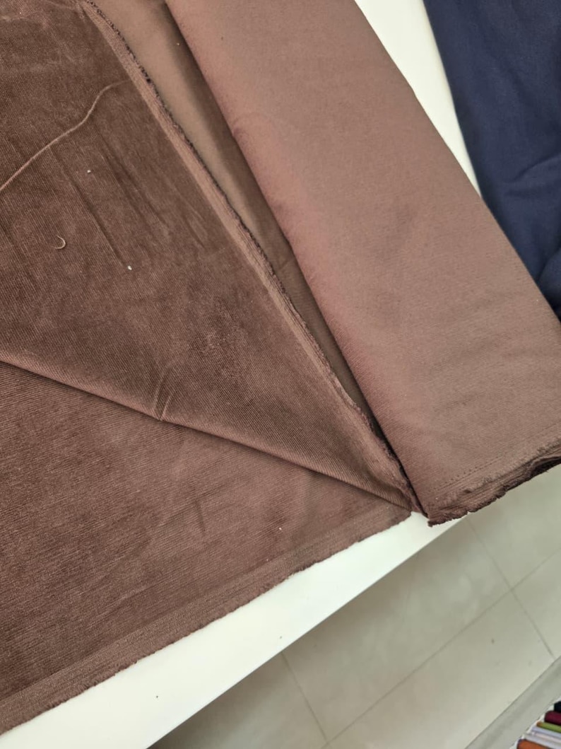 Pu&ograve; includere: Un rotolo di tessuto in velluto a coste marrone caratterizzato da una distinta trama a coste verticali. Il materiale &egrave; ripiegato per rivelare la superficie morbida e opaca del tessuto, mostrando il colore marrone profondo e la trama tessile.