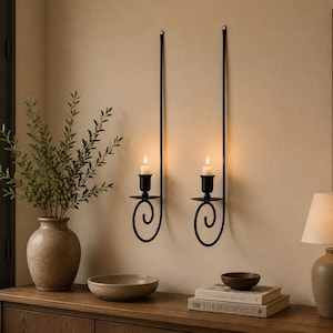 May include: Two black metal wall-mounted candle holders with long, straight vertical stems and a decorative spiral base. Each holder features a small circular platform supporting a lit white pillar candle, creating a symmetrical display.