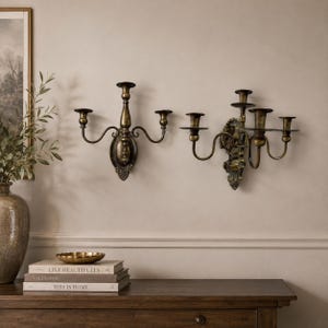 May include: Two antique-style brass wall sconces mounted on a wall. The left sconce features three candle holders with curved arms, while the right sconce has four candle holders with a decorative backplate and intricate metal detailing.