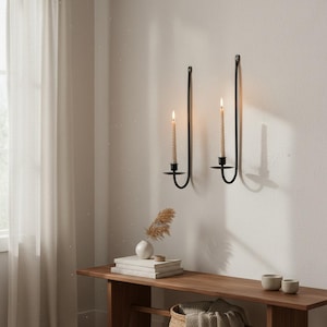 May include: Two black metal wall-mounted candle holders with long, curved arms, each holding a single thin, cream-coloured taper candle. The holders are mounted vertically on a plain wall, with the candles lit, casting a soft glow.