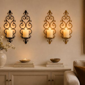 May include: Four wall-mounted candle sconces with ornate scrollwork designs. Two sconces are finished in matte black, and two are finished in a metallic gold tone. Each sconce holds a single cylindrical white pillar candle with a warm glow.