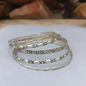 Può includere: Un set di cinque bracciali rigidi in metallo color argento impilati. I bracciali presentano texture varie tra cui bordi ondulati lisci, un motivo a foglie ripetuto, un dettaglio floreale e un design a perline con piccoli accenti circolari.