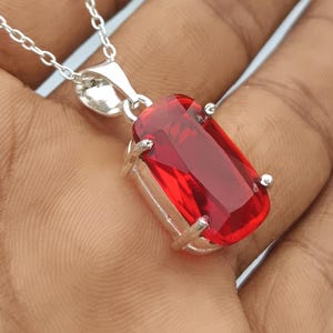Puede incluir: Un colgante de piedra preciosa rectangular, facetada y de color rojo vibrante, montado en un engaste de metal plateado pulido. El colgante cuelga de una delicada cadena de eslabones plateados, mostrando un color rojo intenso y brillante.